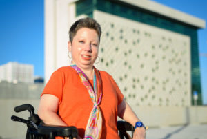 Photo of author Heidi Johnson-Wright, wearing a red, short-sleeved top, a colorful scarf and hoop earrings. She's sitting in a wheelchair, smiling.