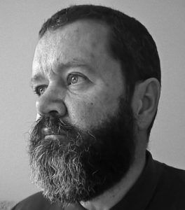 A black and white profile photo of Oleb Books author Nigel David Kelly, a man in his mid-50s with a full, well-trimmed beard.