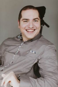 Oleb Books author Andrew Gurza is sitting in his wheelchair, wearing a grey dress shirt, smiling at the camera.