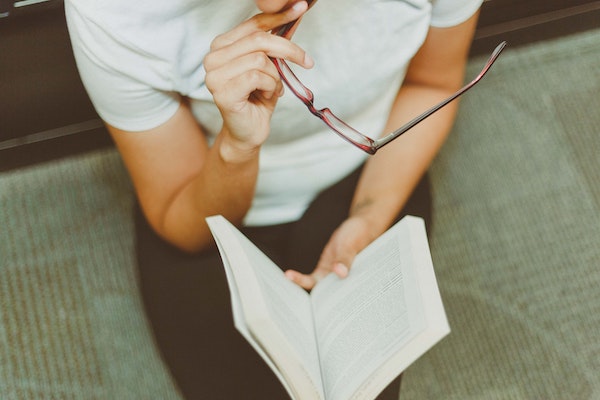 A photo of a man holding a pair of glasses, thoughtfully looking at an open book in his lap.