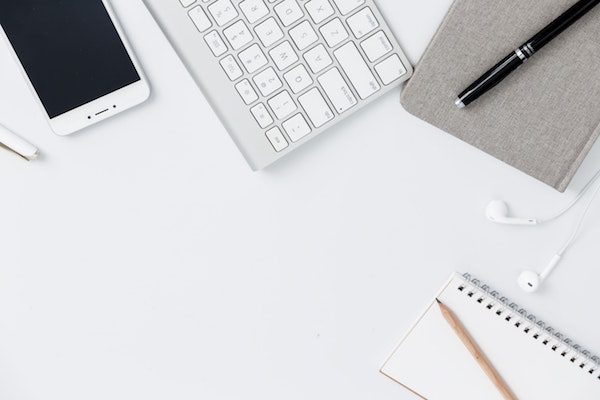 A smartphone, keyboard and notepads and pens, and a set of Apple earbuds are fanned out on a white surface.