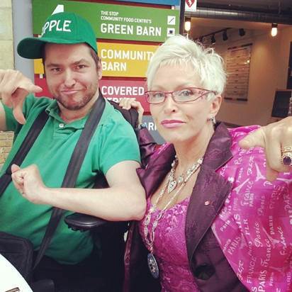 Oleb Books author Andrew Gurza is sitting in a wheelchair wearing a green shirt and hat and is posing happily alongside his mother.