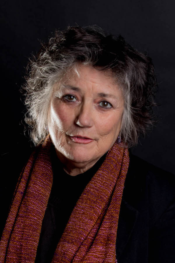 A headshot of author Terry Galloway, with short grey hair in a stylish, messy cut, wearing a black shirt and a red scarf.