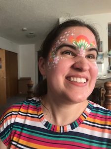 Caitlin Hernandez is beaming as she wears rainbow face paint with stars, dots, and flower petals, and a rainbow dress.