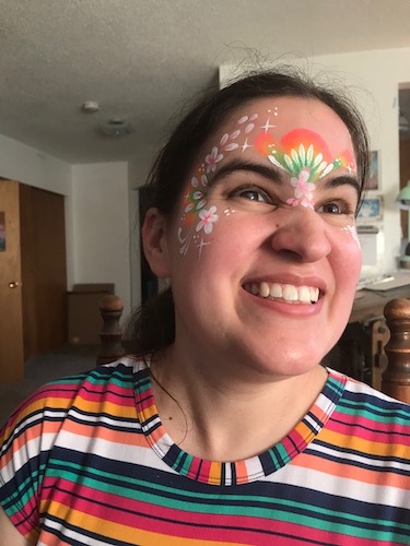 Caitlin Hernandez is beaming as she wears rainbow face paint with stars, dots, and flower petals, and a rainbow dress.
