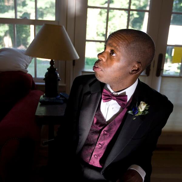 Leroy Moore sits, looking off to the side, wearing a shiny black tux jacket, with purple vest and bowtie.