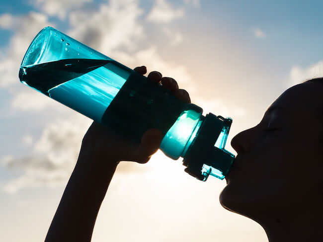 A silhouette of a person drinking water from a bight blue water bottle with a bright sky in the background.
