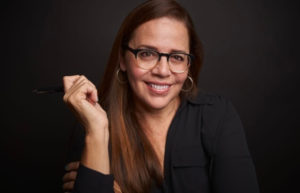 Reine Dugas smiling, wearing a black shirt, hoop earrings and black-rimmed glasses.