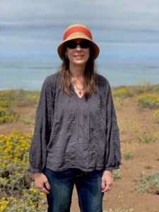 Author Wendy Kennar stands outside smiling, wearing a hat and sunglasses, with the ocean behind her.