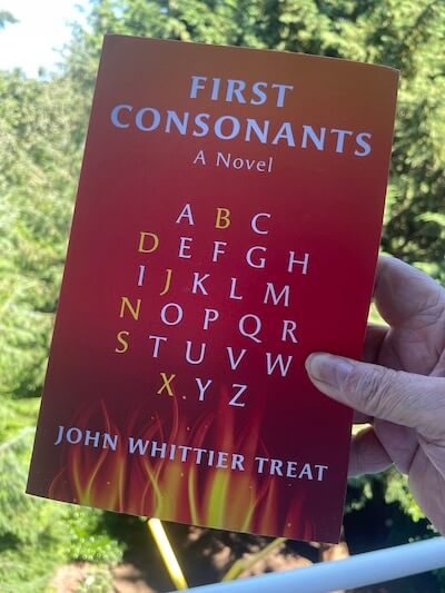 A hand holding up the book First Consonants by John Whittier Treat on a sunny day outdoors.